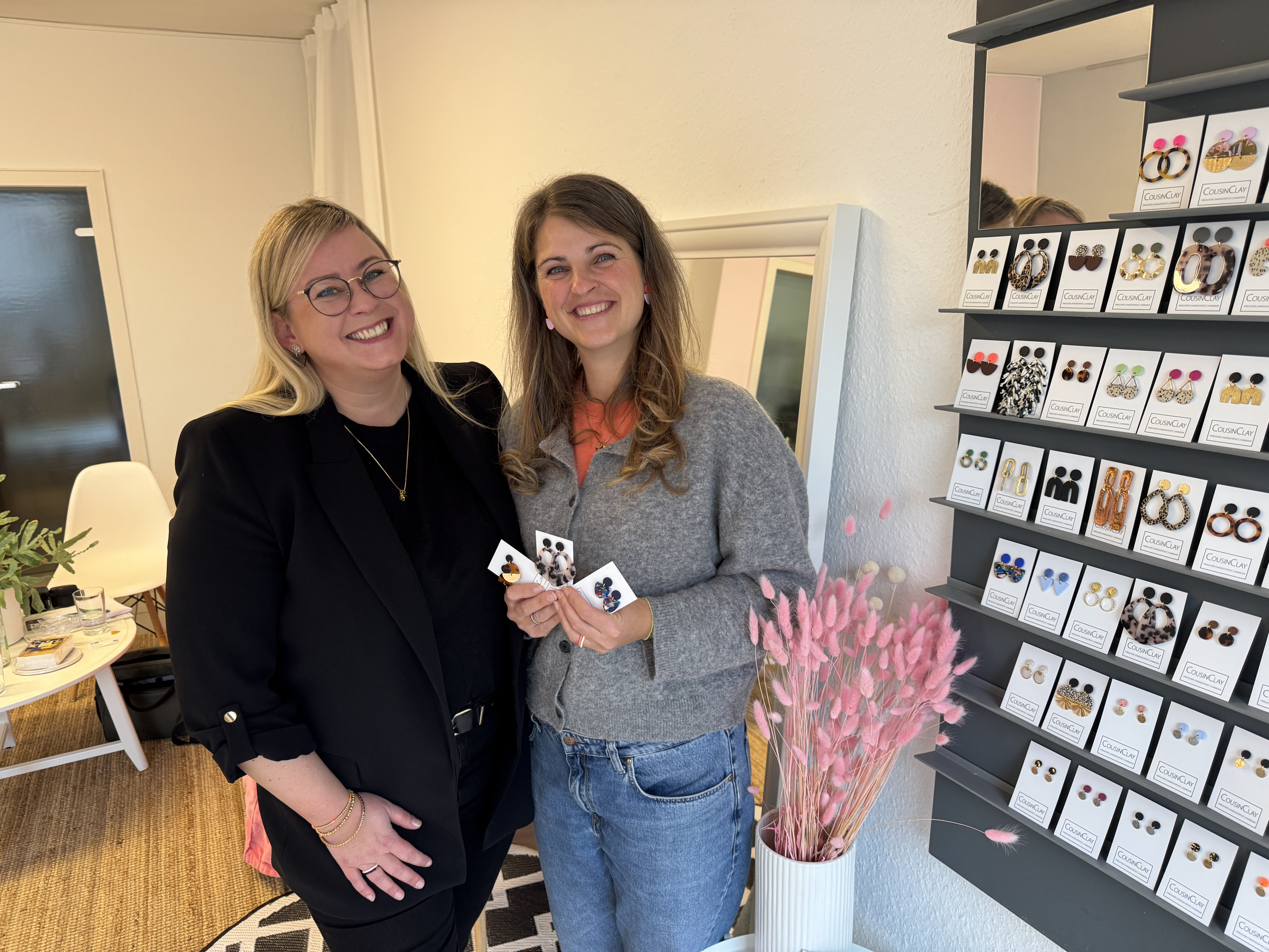 Zwei Frauen in einem Schmuckgeschäft halten Ohrringe in der Hand, im Hintergrund ein Regal mit verschiedenen Schmuckstücken und eine Vase mit rosa Trockenblumen.