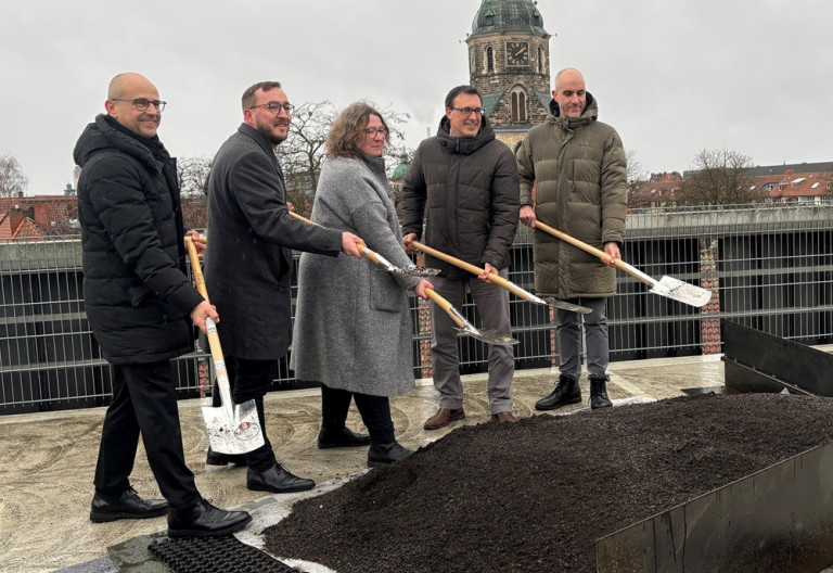 Spatenstich auf einem Dach mit mehreren Personen und Kirchturm im Hintergrund