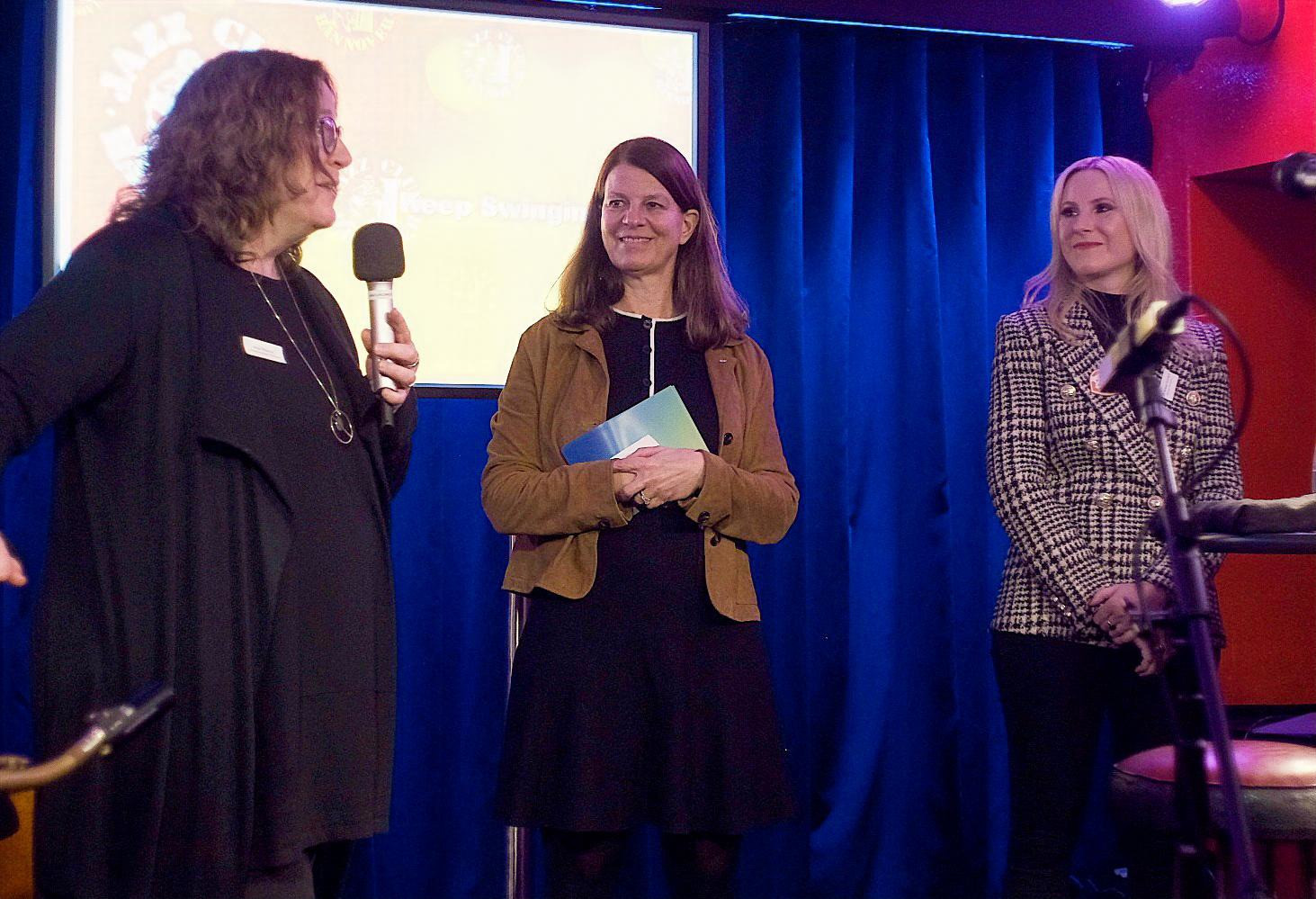 Personen auf einer Veranstaltung mit Mikrofon und Unterlagen vor blauem Vorhang