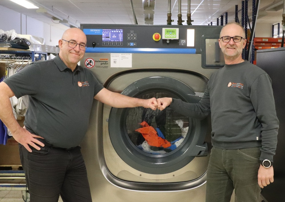 Zwei Personen geben sich einen Fistbump vor einer industriellen Waschmaschine in einer Wäscherei