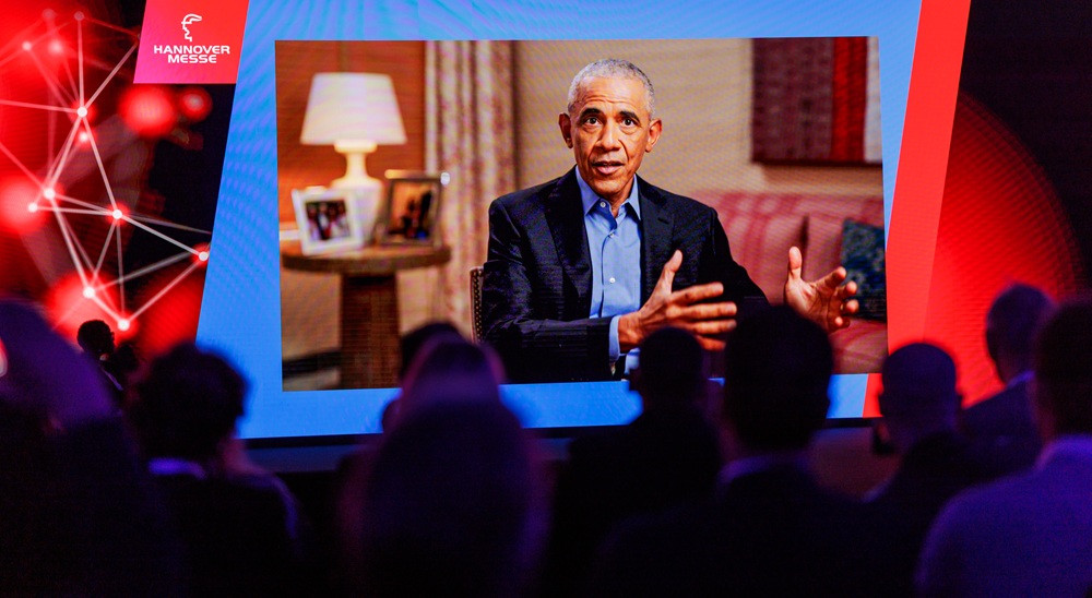 Barack Obama bei einer Videokonferenz-Schalte auf einer Leinwand vor Publikum