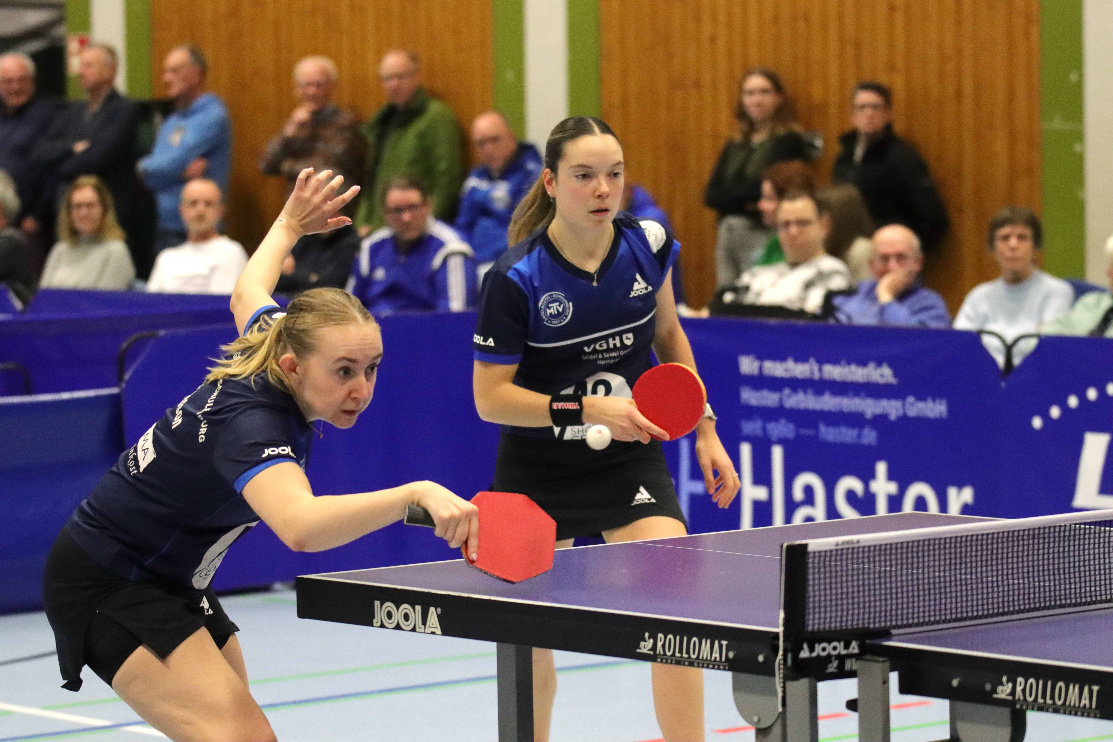 Zwei Tischtennisspielerinnen beim Doppel im Wettkampf, Fokus auf den Aufschlag, Sporthalle mit Zuschauern im Hintergrund