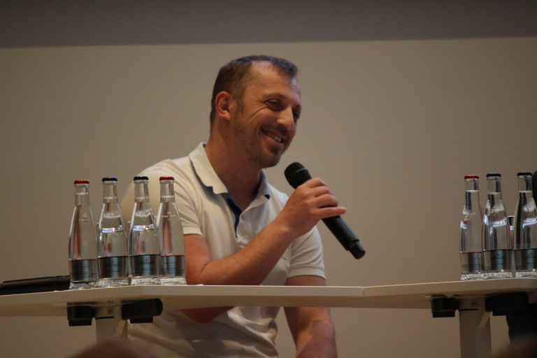 Mann mit Mikrofon bei Podiumsdiskussion, img-1151.JPG Mann im weißen Polohemd spricht mit Mikrofon auf Podium, drei Wasserflaschen auf Tisch