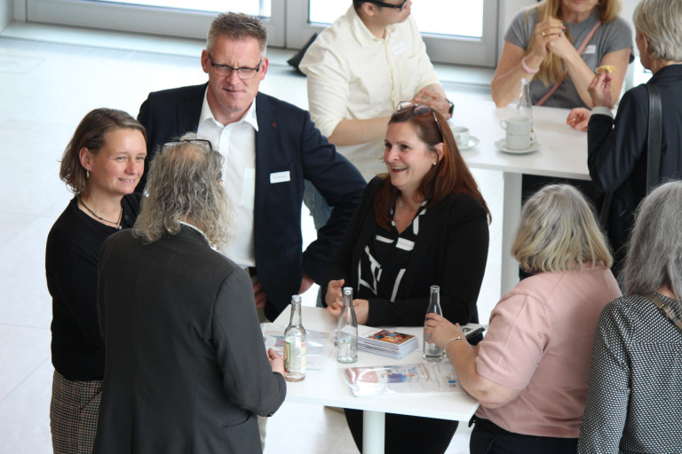Networking-Veranstaltung: Personen im Gesprch an einem Stehtisch mit Unterlagen und Getrnken Mehrere Personen im Gesprch bei einer Networking-Veranstaltung an einem Stehtisch mit Unterlagen und Getrnken