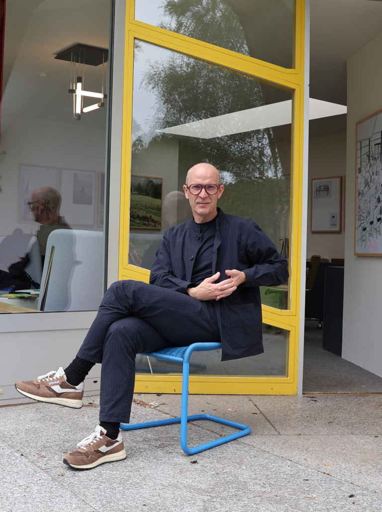 Mann mit Brille sitzt entspannt auf einem blauen Stuhl vor einem modernen Fenster mit gelbem Rahmen