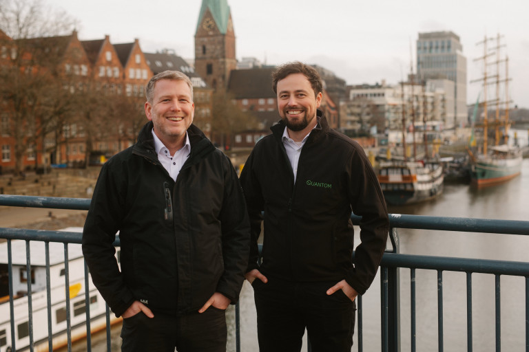 Zwei Männer auf einer Brücke mit maritimer Kulisse in der Stadt, einer trägt eine Jacke mit dem Schriftzug QUANTOM.