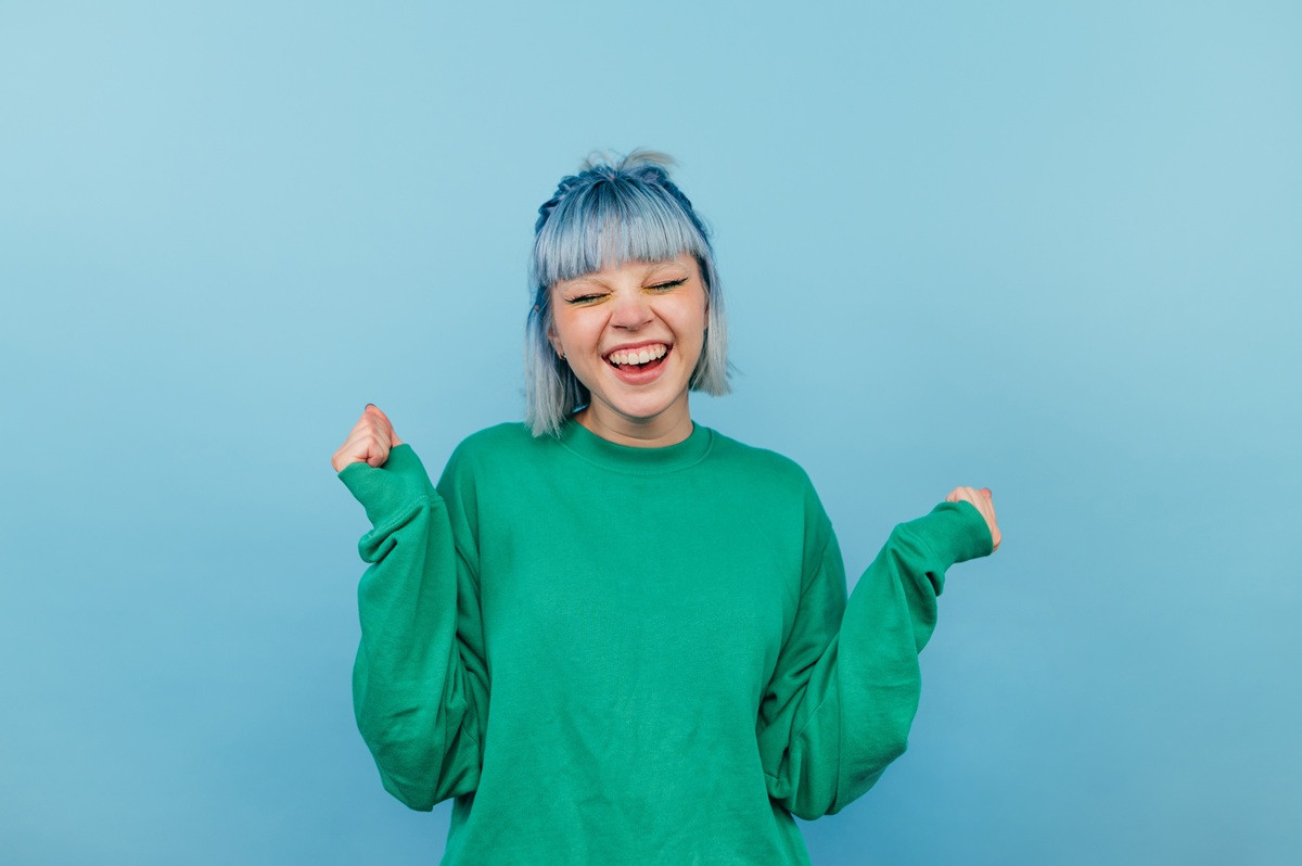 adobestock-494324869-web: Glückliche junge Frau mit blauen Haaren und grünem Pullover vor blauem Hintergrund