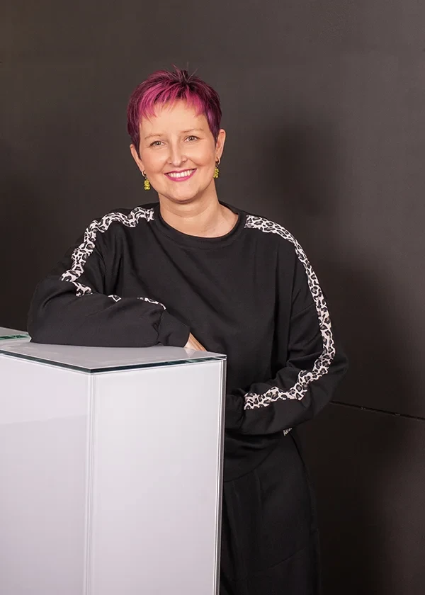 Olga mit kurzem pinkem Haar, lächelnd vor dunklem Hintergrund