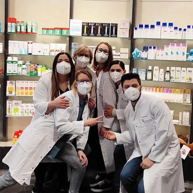 Teamfoto von Apothekenmitarbeitern mit weißen Kitteln und Masken vor einem Regal mit Medikamenten