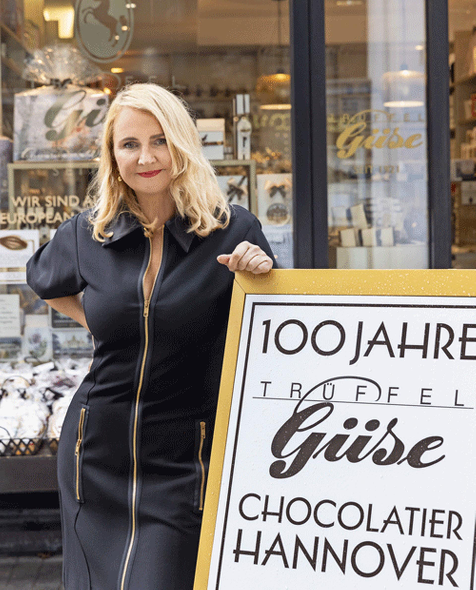 Vor dem Laden in Hannovers Innenstadt: Der Jubiläumsaufsteller macht auf 100 Jahre Trüffel-Genuss aufmerksam. Chefin Sabine Güse-Henschel steht vor dem Eingang der Schokoladenmanufaktur, neben einem Aufsteller mit Hinweis auf das 100-jährige Jubiläum.