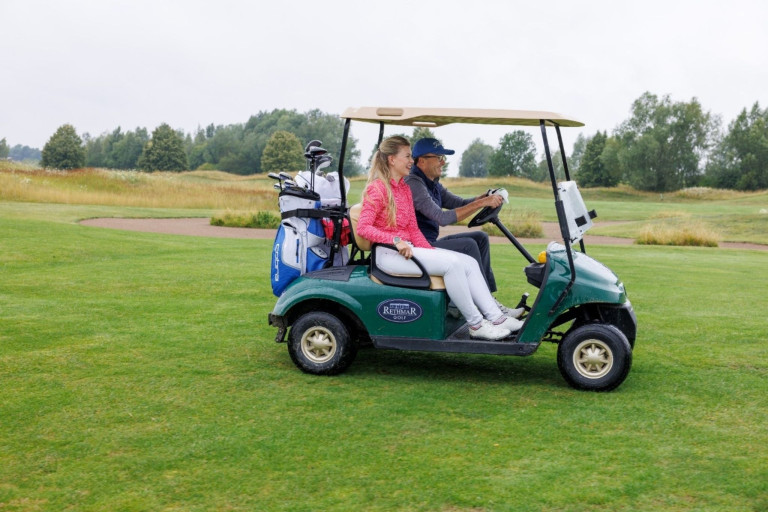 Zwei Personen fahren mit einem Golfcart auf dem Golfplatz Rehthar, Golfbags im Hintergrund, grüne Wiese und Bäume