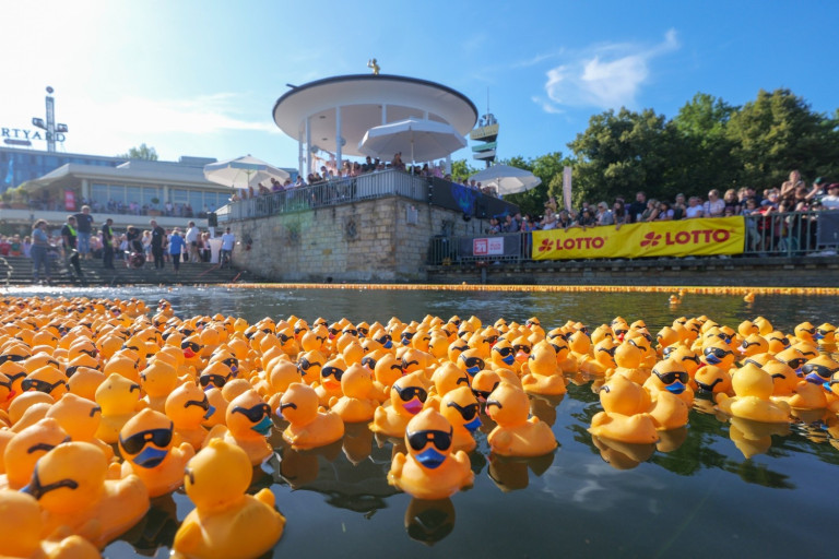 Viele gelbe Gummienten mit Sonnenbrillen beim Entenrennen auf einem Fluss, im Hintergrund Zuschauer und ein Lotto-Banner.