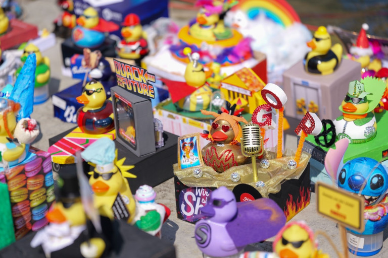 Bunte, kreativ dekorierte Gummienten auf einer Ausstellung, unter anderem mit Mikrofon, Sonnenbrille und Motto-Schildern wie 'Quack to the Future'.