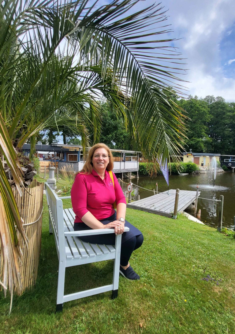 Sonja de Vries sitzt auf einer weißen Bank im Grünen an einem Teich mit Steg und Palmen