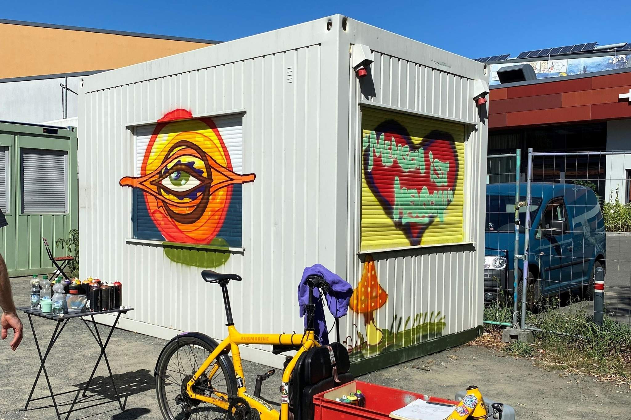 Graffiti-Kunst auf Container: Auge, Herz mit 'Mensch ist Mensch' und gelbes Fahrrad Graffiti auf weißem Container mit buntem Auge, Herz und Schriftzug 'Mensch ist Mensch', davor gelbes Fahrrad