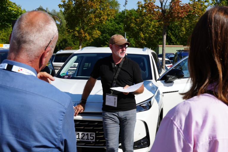 Vortrag beim Forum Mobility Work Event im Freien vor einem weißen Fahrzeug Person hält einen Vortrag vor Teilnehmern bei einem Forum Mobility Work Event im Freien vor einem weißen Auto