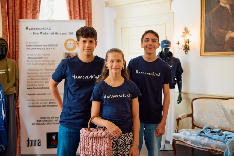 Hannoverkind T-Shirts und Herabag Tasche auf Event 2025 von Karsten Koch Drei Jugendliche tragen Hannoverkind T-Shirts und halten eine Herabag Tasche, aufgenommen bei einem Event 2025 von Karsten Koch.