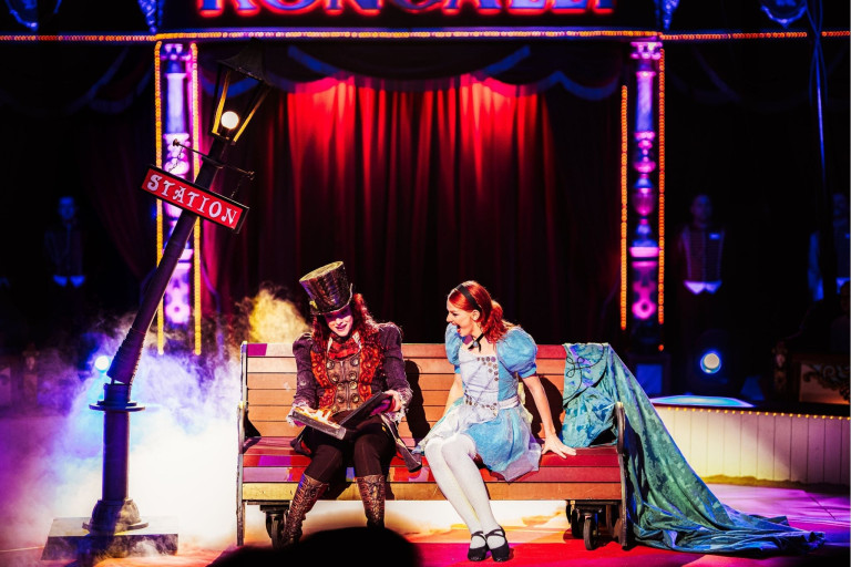 Roncalli Zirkus Szene mit Darstellern auf der Bühne Roncalli Zirkus Show mit zwei Darstellern auf einer Bank vor rotem Vorhang