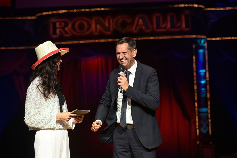 Zwei Personen auf der Bühne beim Circus Roncalli, einer hält ein Mikrofon, im Hintergrund das Roncalli-Logo