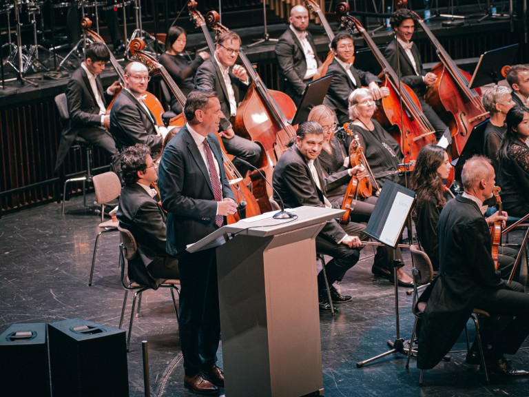 Peer Bothmer bei Freude HCC Event mit Orchester auf der Bhne Peer Bothmer spricht bei einer Veranstaltung mit Orchester auf der Bhne, Freude HCC Event