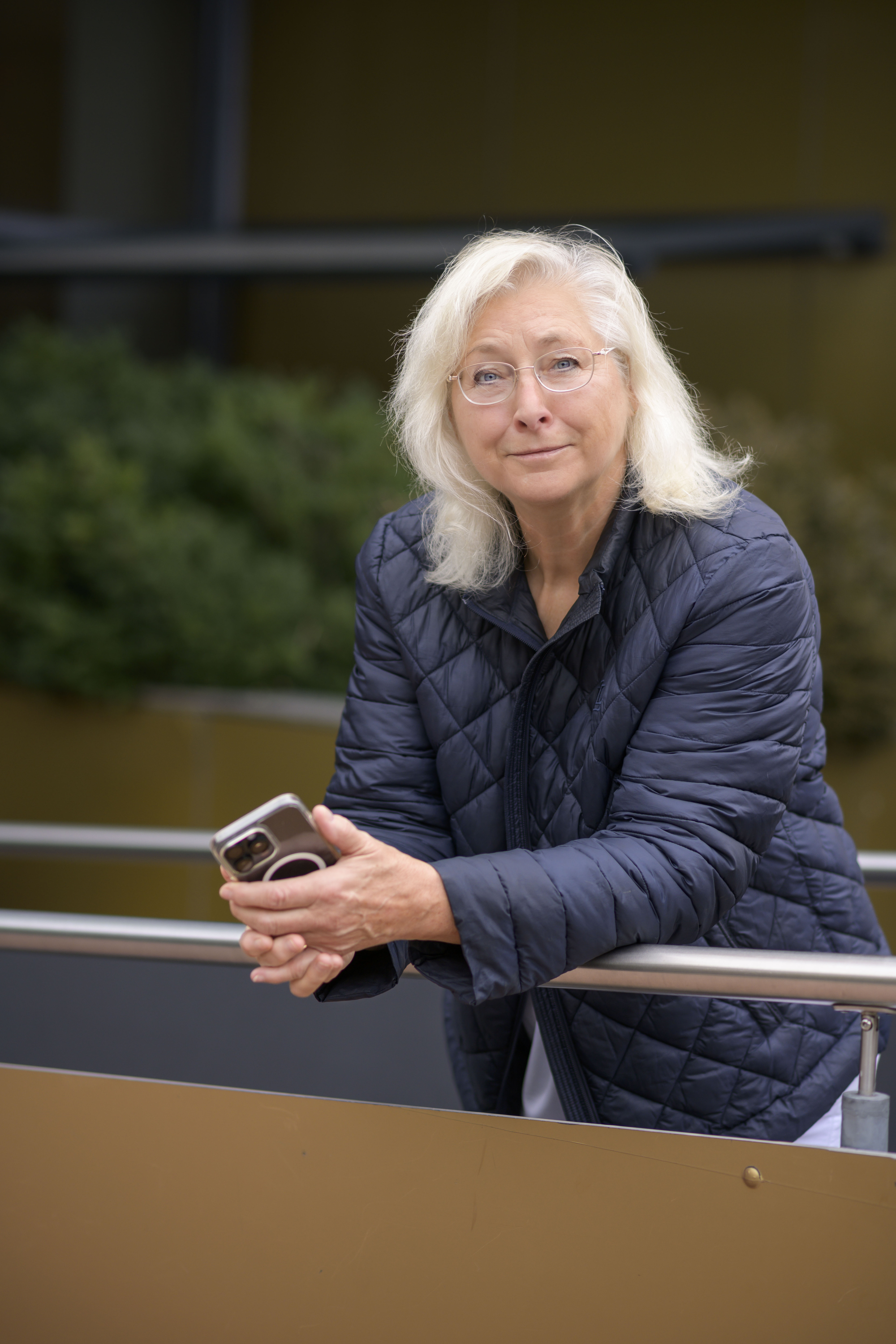 Ältere Frau mit weißem Haar und blauer Steppjacke hält ein Smartphone in der Hand und lehnt an einem Geländer im Freien.