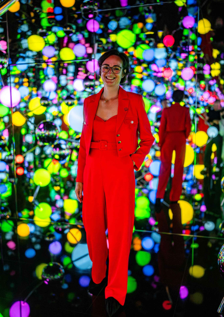 Aurelie in rotem Anzug vor bunter, leuchtender Kunstinstallation