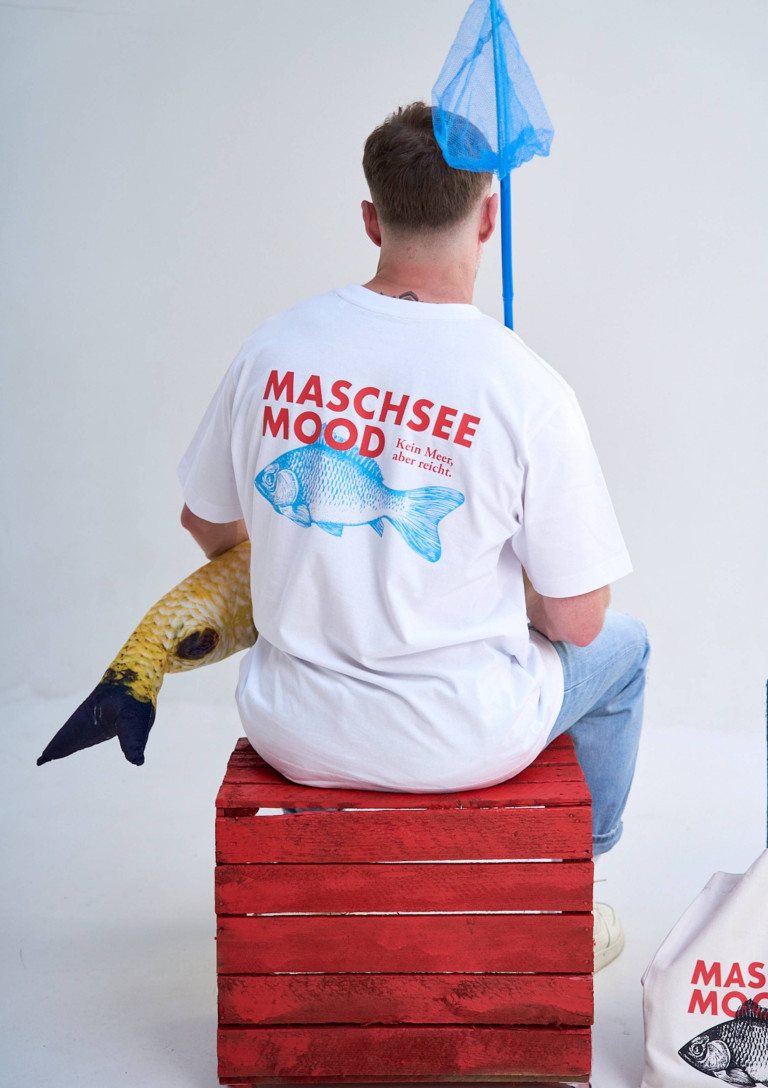 Rückansicht eines Mannes mit weißem T-Shirt mit Aufdruck 'Maschsee Mood', der auf einer roten Kiste sitzt und ein Fisch-Kissen hält.
