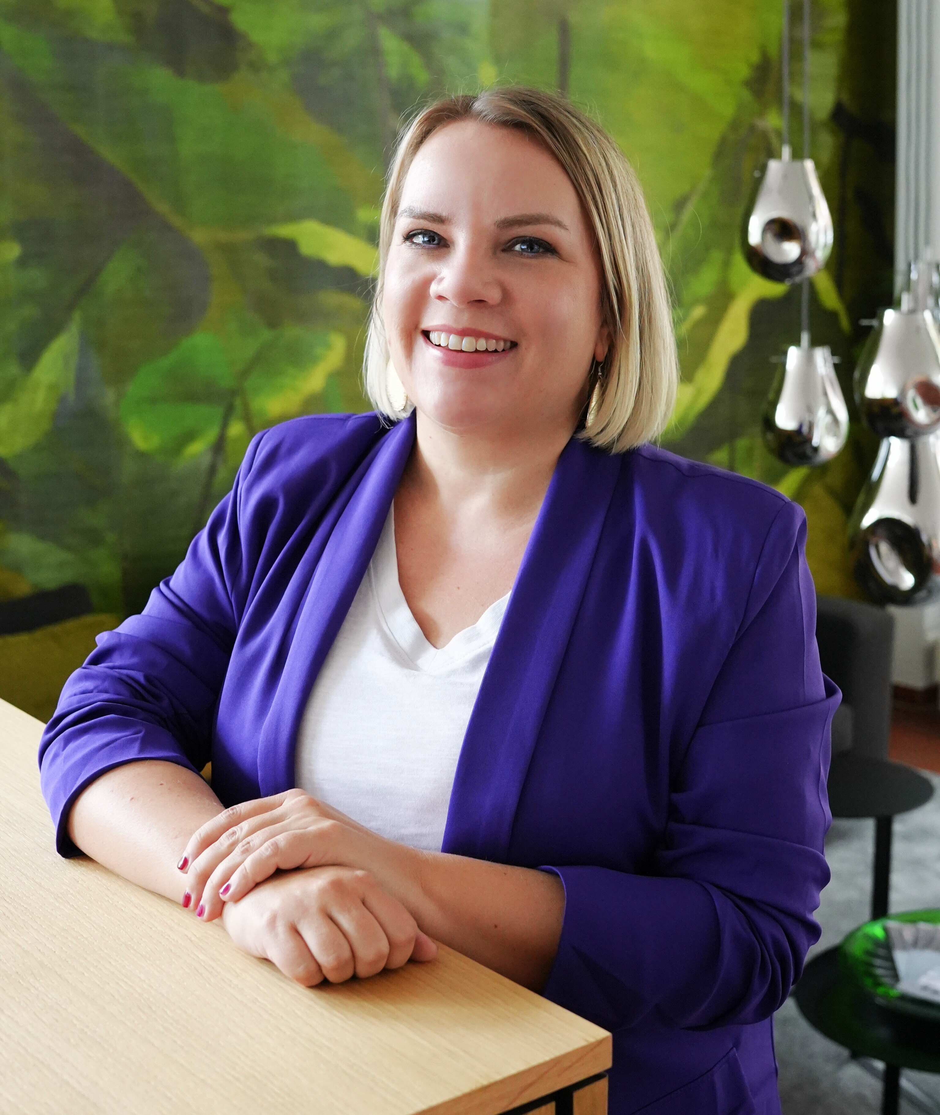 Frau mit blonder Kurzhaarfrisur im lila Blazer sitzt an einem Tisch in modernem Büro