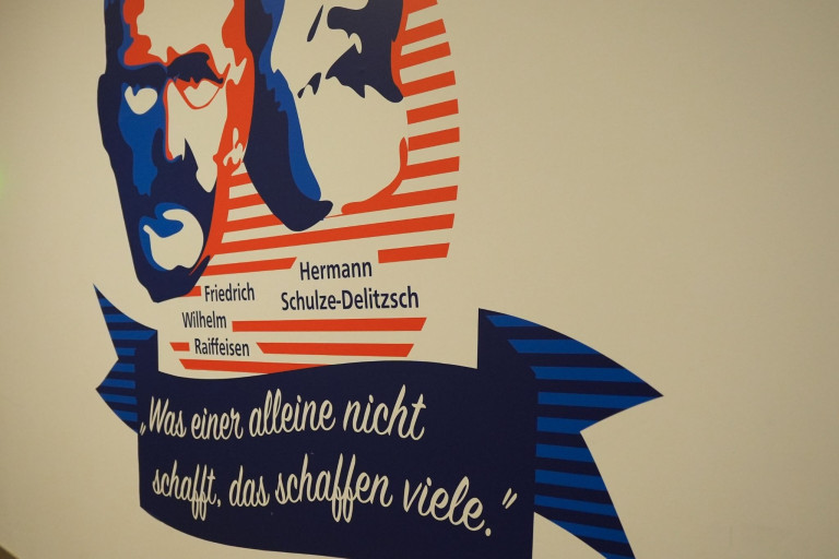 Wandgrafik mit Porträts von Friedrich Wilhelm Raiffeisen und Hermann Schulze-Delitzsch sowie dem Zitat: 'Was einer alleine nicht schafft, das schaffen viele.'