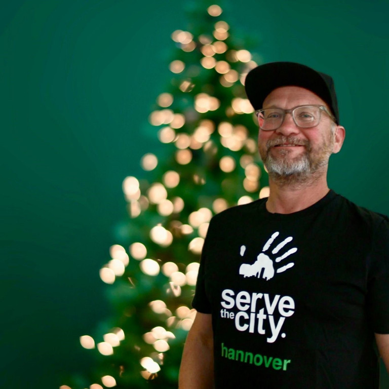 Person mit Serve the City Hannover T-Shirt vor einem unscharfen Weihnachtsbaum beim Mantelhaus Kaiser