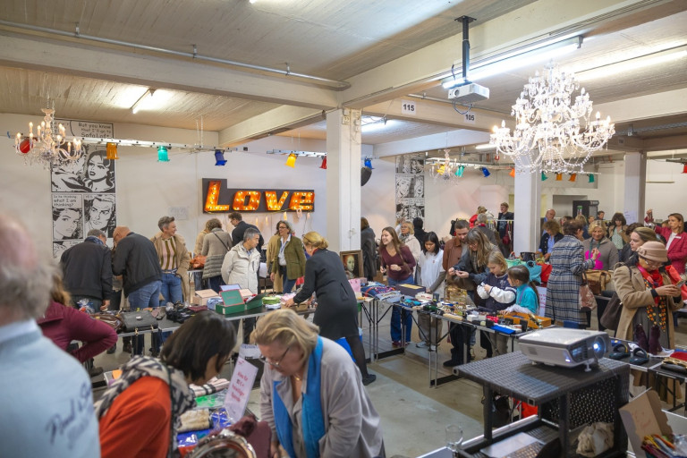 Flohmarkt-Atmosphäre mit Love-Leuchtschild – Foto von Klöpperfotodesign Flohmarkt in einer Halle mit vielen Menschen und einem leuchtenden Love-Schild, fotografiert von Klöpperfotodesign