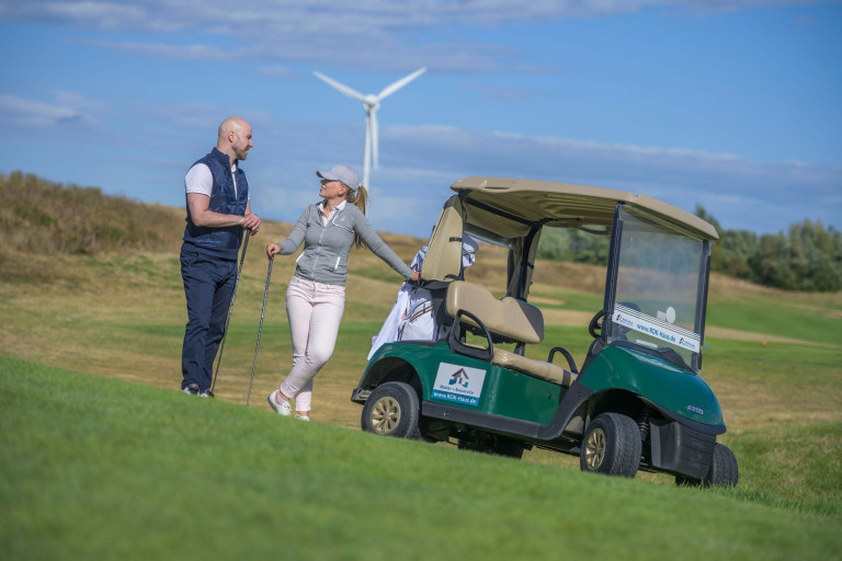 Golfspieler und Golfcart auf dem Golfplatz mit Windrad im Hintergrund Zwei Personen beim Golfspielen neben einem Golfcart auf dem Golfplatz mit Windrad im Hintergrund