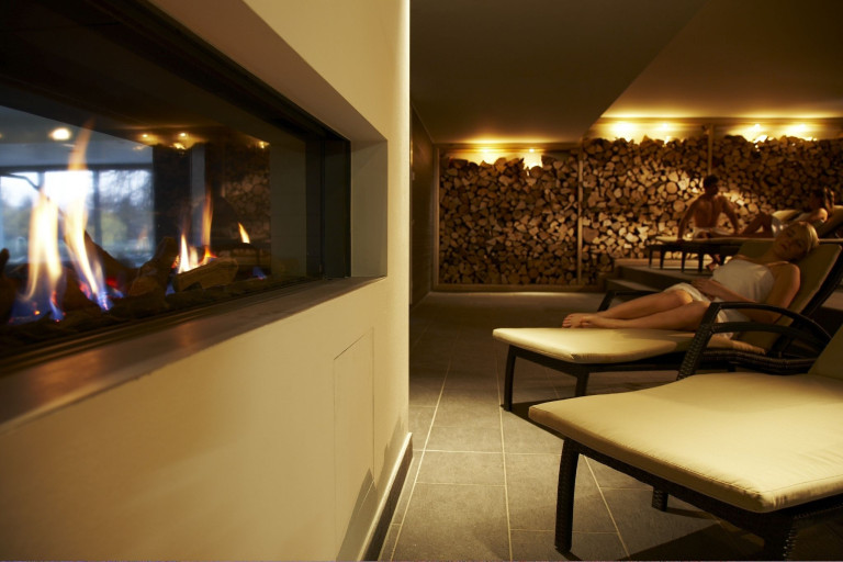 Wellnessbereich mit Kamin im Aspria Club – gemütliche Liegen und Holzstapel im Hintergrund