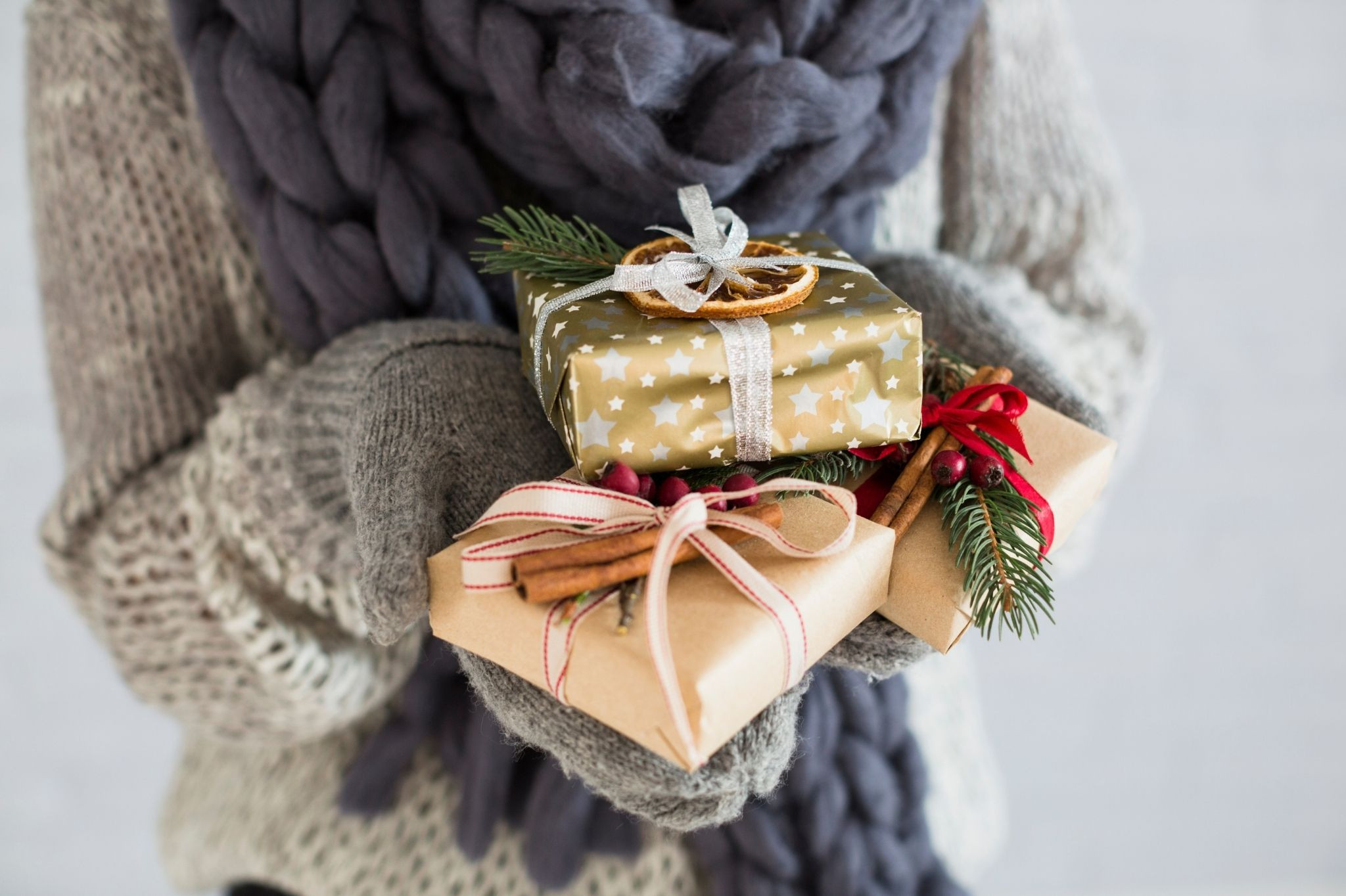 Geschenke in weihnachtlicher Verpackung mit Zimtstangen und Tannenzweigen, gehalten von einer Person mit Wollhandschuhen