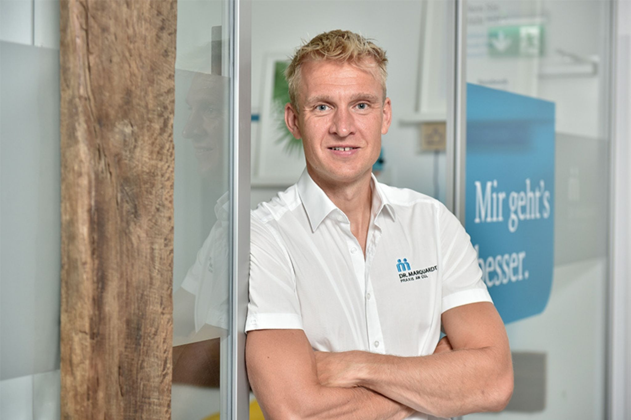 Dr. Matthias Marquardt in weißem Hemd mit Praxislogo, stehend in moderner Praxisumgebung