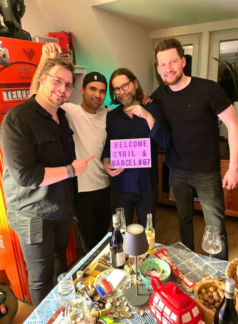 Partybild mit rotem Kühlschrank und Begrüßungsschild für Cyril und Marcel Gruppenfoto bei einer Party mit rotem Kühlschrank und Schild 'Welcome Cyril & Marcel #67'