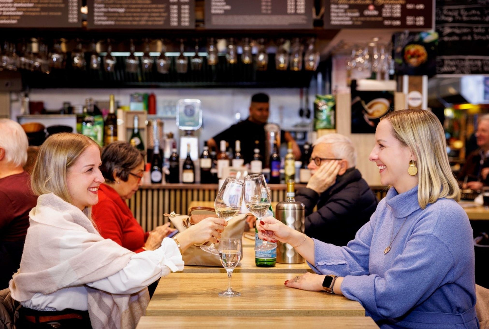 Zwei Frauen stoßen mit Weingläsern in einer Markthalle an, im Hintergrund eine Bar mit Getränken.