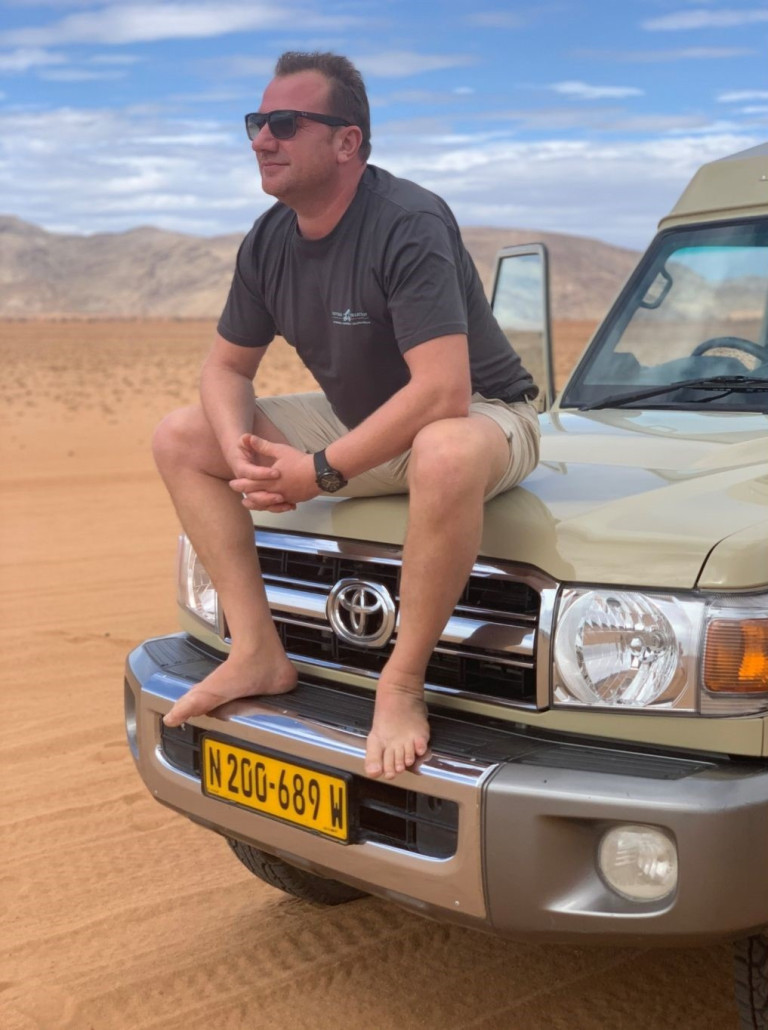 Hans Joachim Bischoff sitzt barfuß auf einem Toyota-Geländewagen in einer Wüstenlandschaft