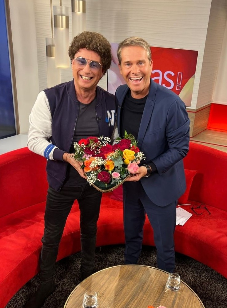 Zwei Personen mit Blumenstrauß in TV-Studio auf rotem Sofa, bilder-nobilis-web