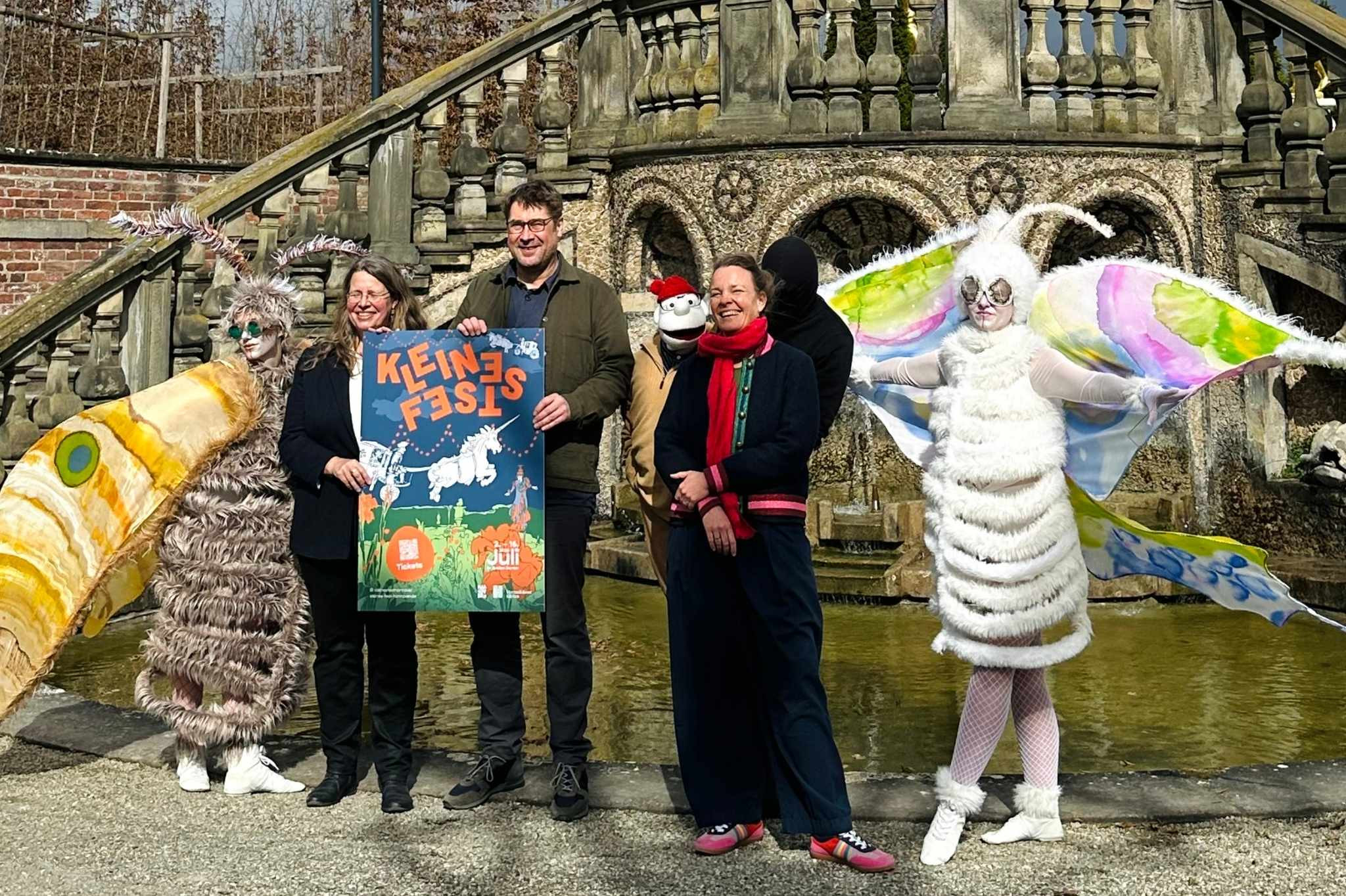 Gruppenfoto mit Kostümierten und Plakat zum Kleines Fest 2026 vor historischer Kulisse