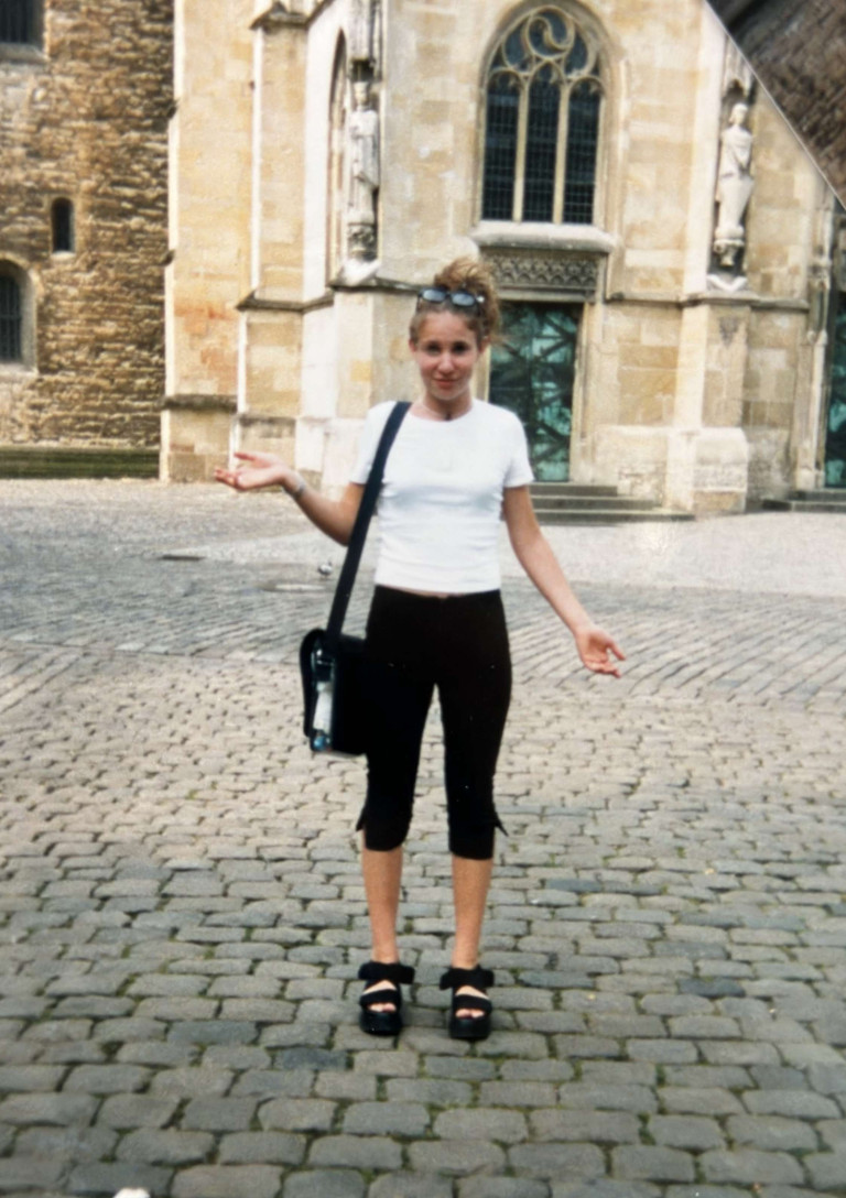 Junge Frau im 90er-Jahre-Outfit auf einem Kopfsteinpflasterplatz vor einem historischen Gebäude