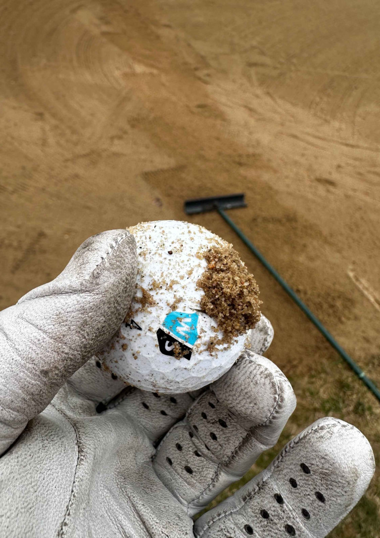 Golfball mit Sand im Bunker, gehalten von einer behandschuhten Hand – grasgefluester-1