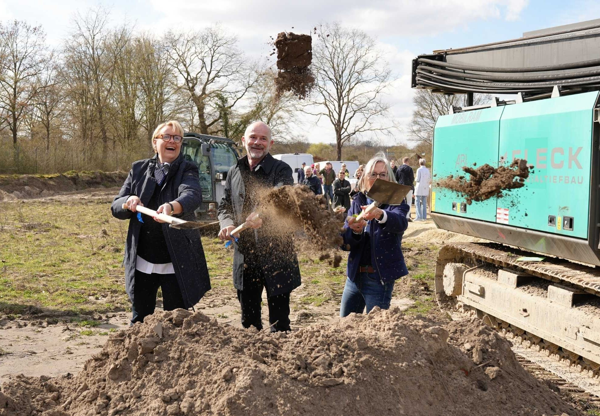 Spatenstich bei einer Baustelle mit mehreren Personen, die Erde mit Schaufeln werfen, im Hintergrund Bagger und Zuschauer