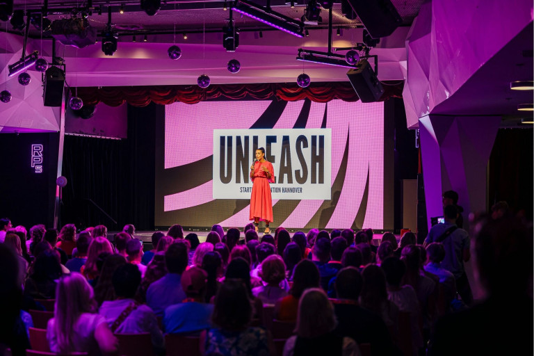 Frau auf der Bühne bei der UNLEASH Startup Convention Hannover, veranstaltet von hannoverimpuls, vor großem Publikum.
