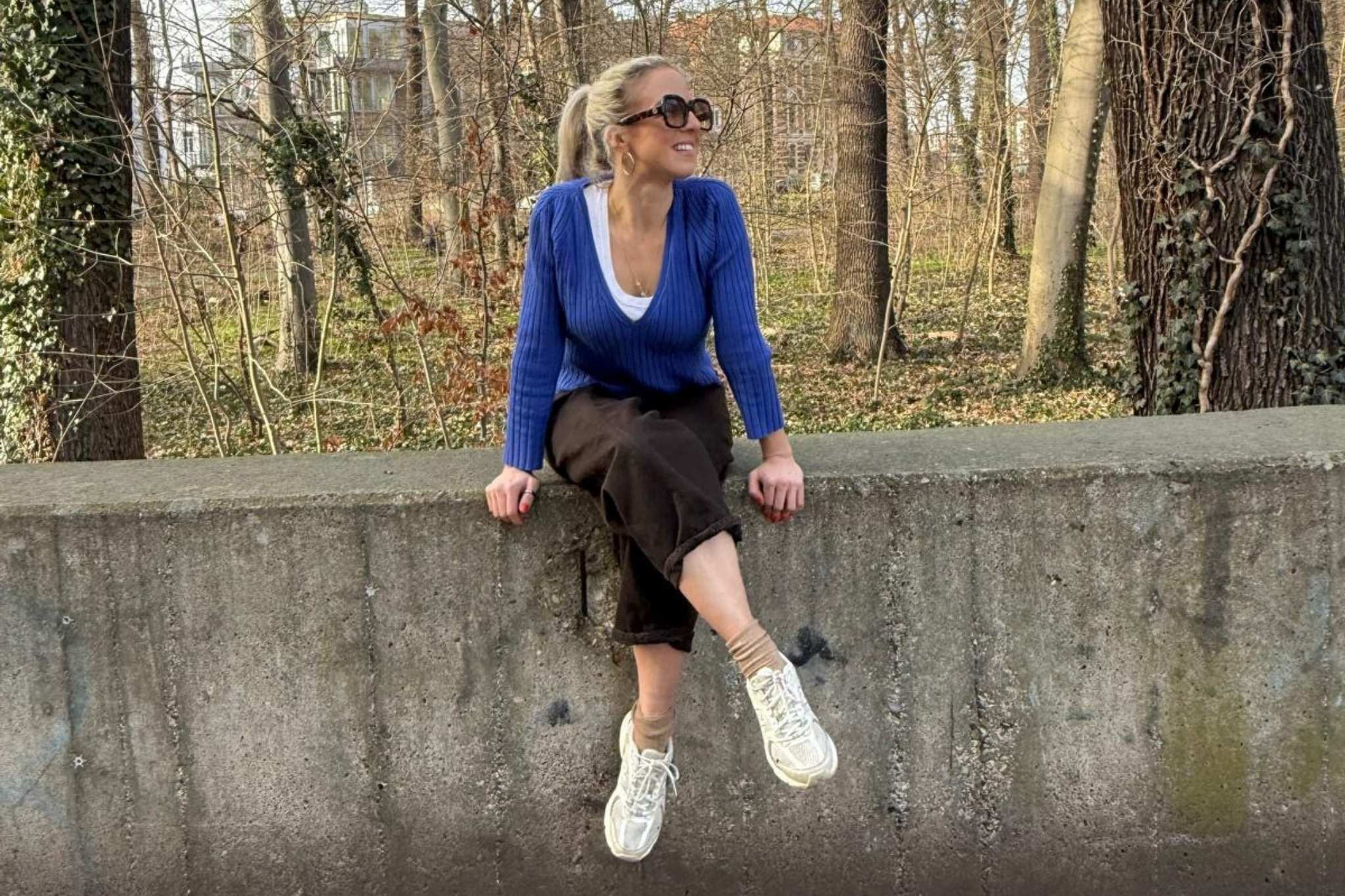 Lara Kertess sitzt entspannt auf einer Betonmauer im Park mit blauem Pullover