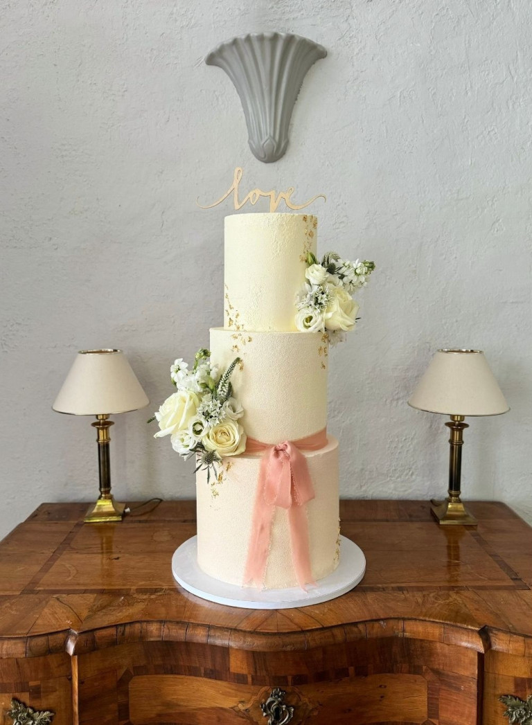 Dreistöckige Hochzeitstorte mit weißen Blumen und rosa Schleife auf Holztisch, dekoriert für eine Hochzeit, aufgenommen für Nobilis Web.