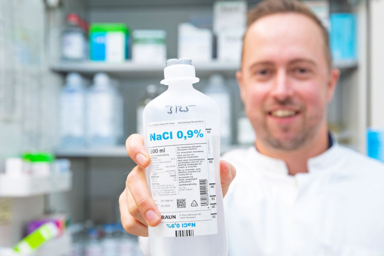 Person hält eine Flasche mit NaCl 0,9% Kochsalzlösung in einer Apotheke oder medizinischen Einrichtung, lachmann-1
