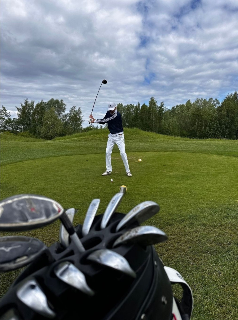 Golfer beim Abschlag auf dem Golfplatz, im Vordergrund Golfbag mit Schlägern, unter bewölktem Himmel