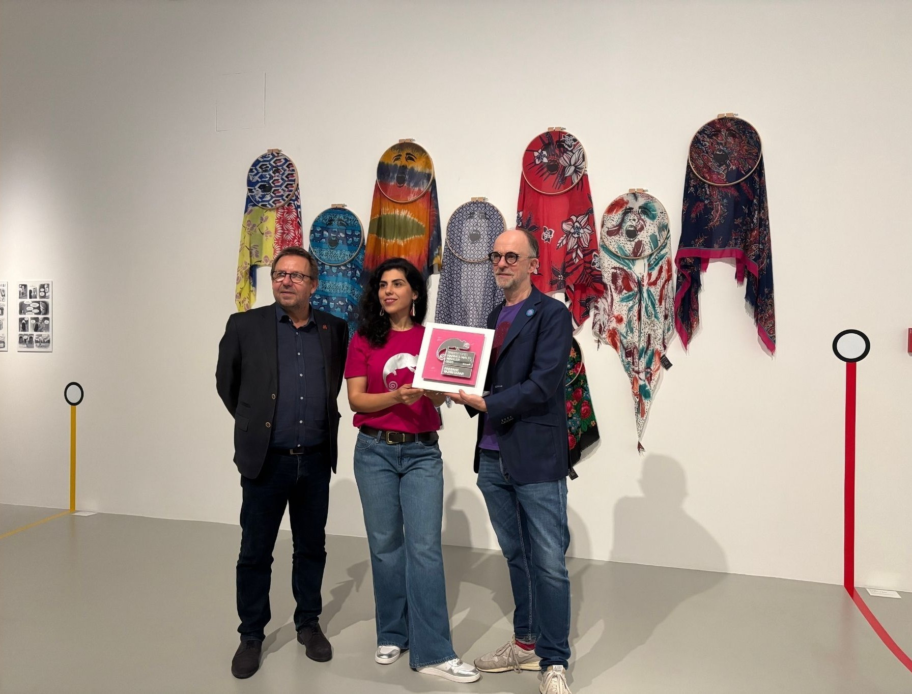 Drei Personen stehen vor einer Wand mit bunten Tüchern und halten eine Auszeichnung, Ausstellung Nobilis