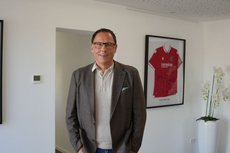 Artikel Michele Hust: Mann vor Hannover 96 Trikot in modernem Büro Mann im Sakko vor signiertem Hannover 96 Trikot – Artikel Michele Hust